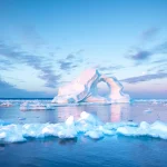 Photogenic and intricate iceberg with a hole under an interesting and colorful sky during sunrise with full moon. Disko bay, Greenland.