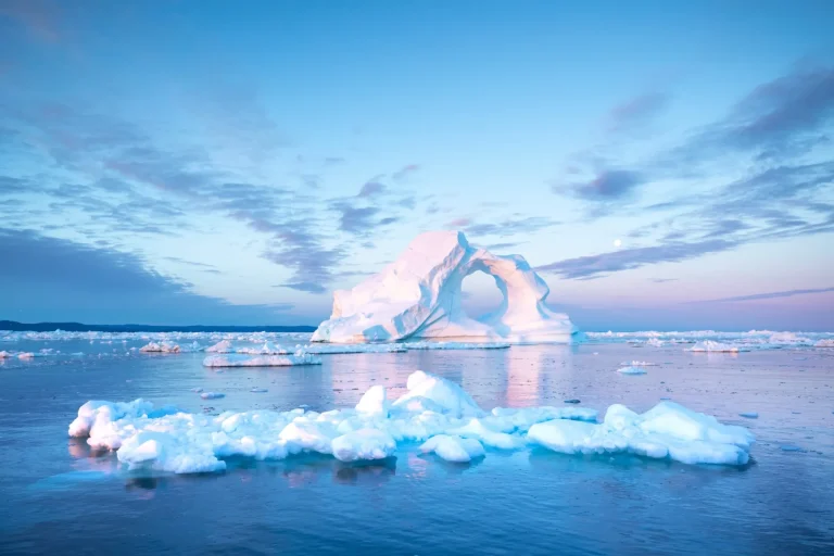 Photogenic and intricate iceberg with a hole under an interesting and colorful sky during sunrise with full moon. Disko bay, Greenland.
