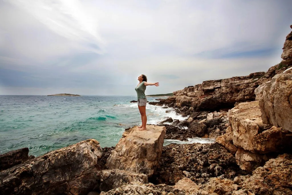 Woman with outstretched arms enjoying the wind and breathing fresh air on the rocky beach
